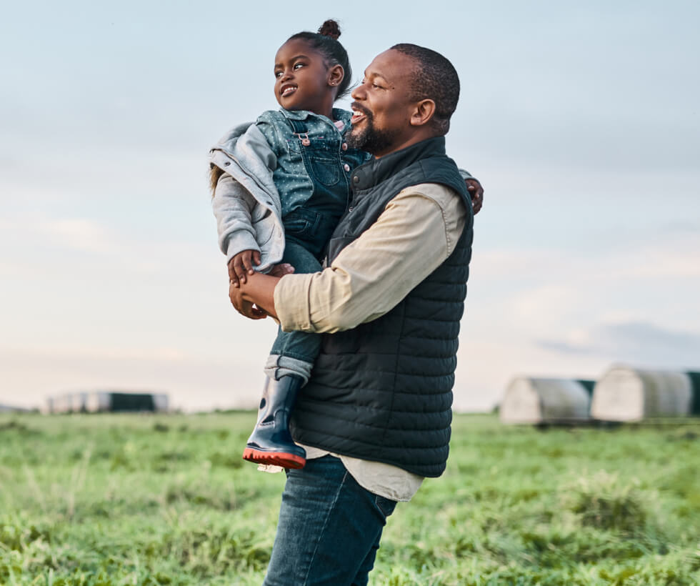 A dad carrying a small child through a field with farm buildings in the background